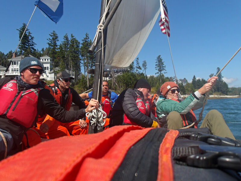 A group of people are on a sailboat, possibly racing or enjoying a leisurely sail. They are wearing life jackets, and some are holding ropes, indicating they are actively involved in sailing the boat. The sky is clear and blue, suggesting a sunny day. The presence of trees on the coastline indicates they are sailing near land.